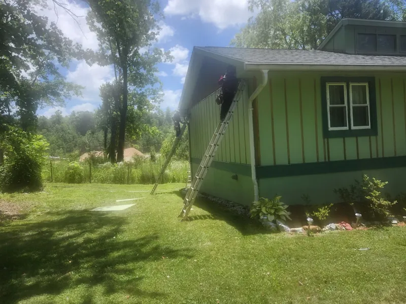 Ladders set against a green board-and-batten house with a crew member at the roofline
