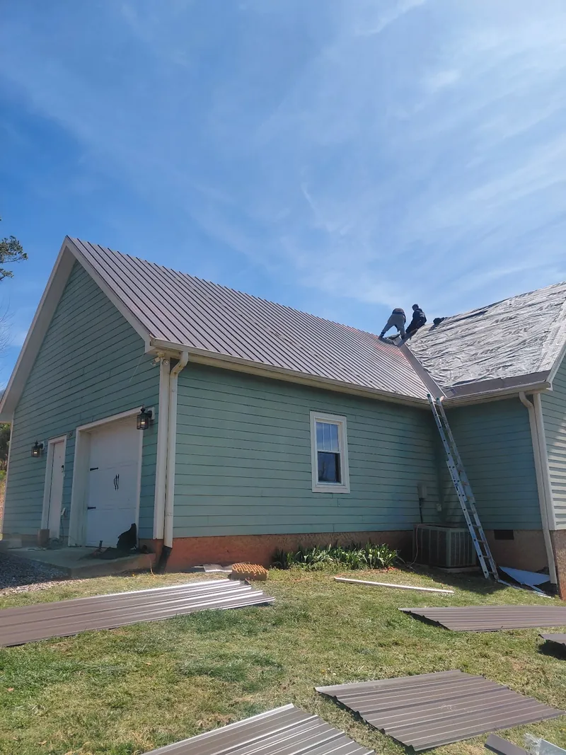 A standing-seam metal roof going onto a light blue house, with a crew member at the ridge and panels stacked on the lawn